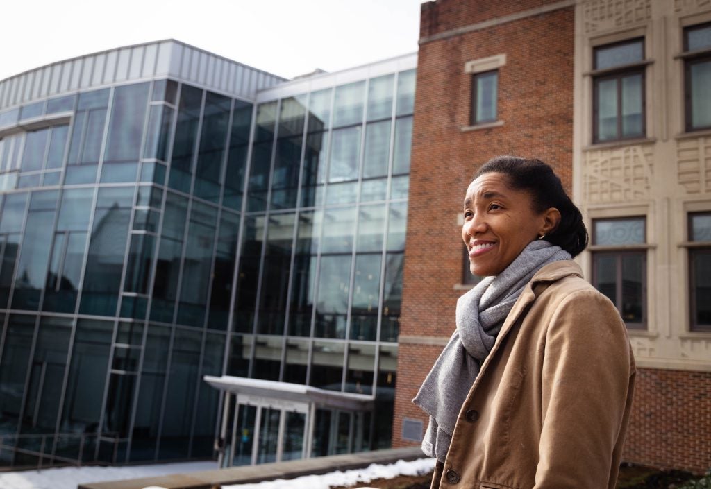 Una mujer negra con una chaqueta color canela y una bufanda gris sonríe frente a un edificio moderno en el campus de Georgetown.