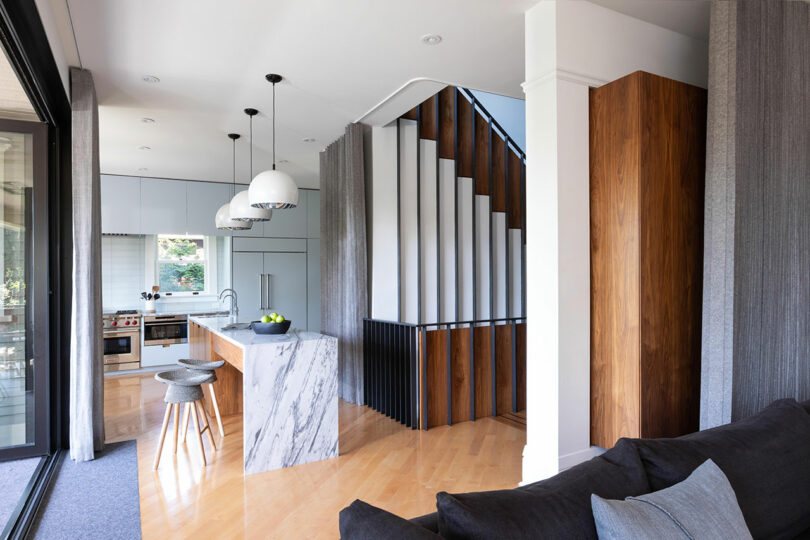 Una cocina moderna con isla de mármol junto a un salón con un sofá gris y una escalera con paneles verticales de madera y blancos.