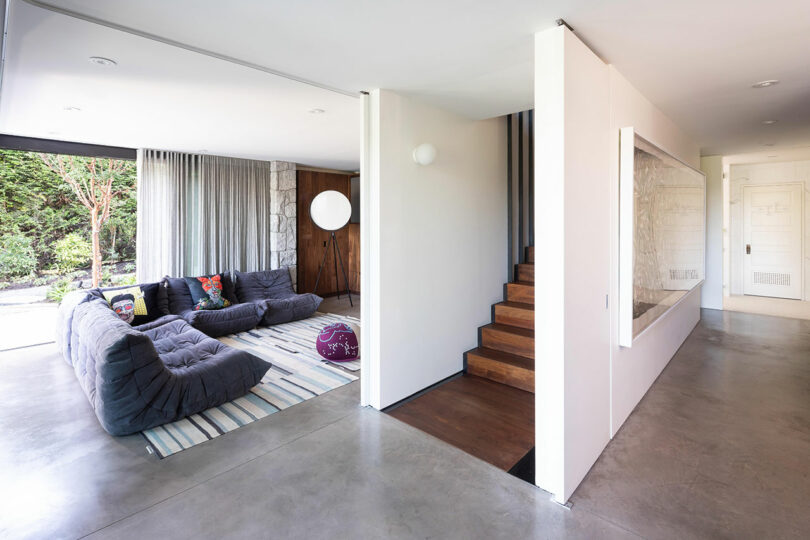 Una sala de estar espaciosa y moderna con un gran sofá seccional gris, escaleras detrás de una pared parcial y ventanas del piso al techo que muestran la vegetación del exterior.