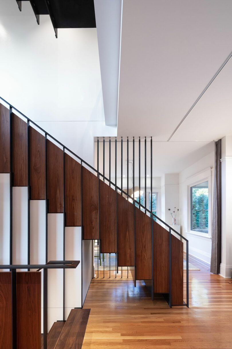 Escalera moderna en una habitación luminosa con cálidos paneles de madera y barandillas de metal negro, complementadas con elegantes elementos decorativos blancos debajo de las escaleras.