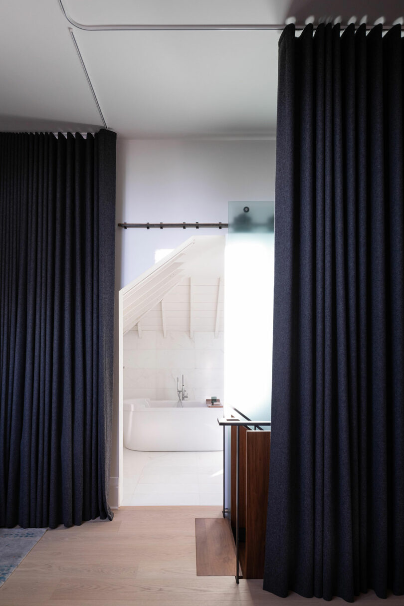El moderno cuarto de baño, visible a través de cortinas opacas, tiene paredes de mármol, una bañera vista y un mueble de madera.