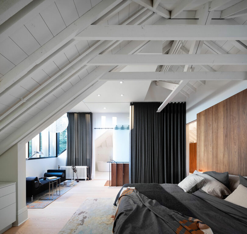 El moderno dormitorio tipo loft tiene vigas blancas, paneles de madera en las paredes, una cama grande y cortinas negras que dividen el espacio.