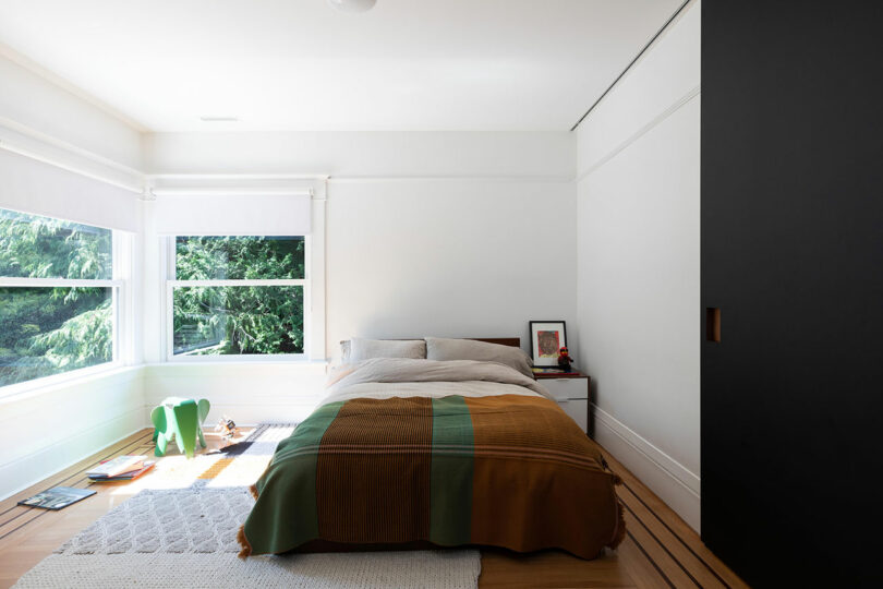 Un dormitorio minimalista con una cama cubierta con una manta verde y marrón, una mesita con libros y un gran ventanal con árboles.