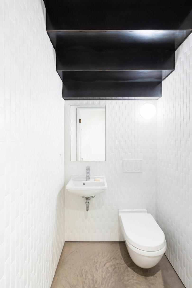 Cuarto de baño pequeño y moderno diseñado con paredes de textura blanca, lavabo de pared, inodoro y techo negro.  Se ven un único espejo y una luz del techo.