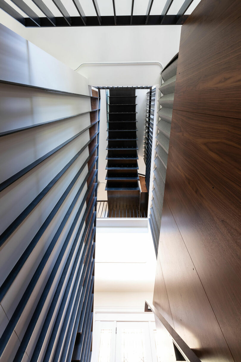 Desde la parte inferior de una escalera moderna con una escalera oscura y medida con paneles de madera y barandillas de metal, mirando hacia un tragaluz brillante.