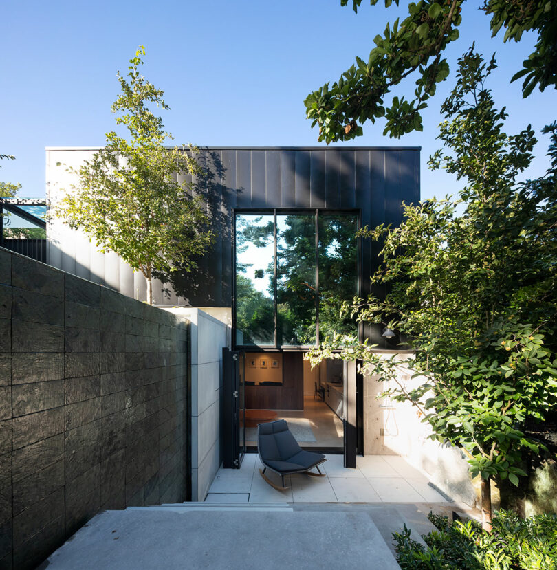 Una casa moderna de dos pisos con grandes ventanales de vidrio, rodeada de exuberante vegetación y una pequeña zona para sentarse al aire libre.