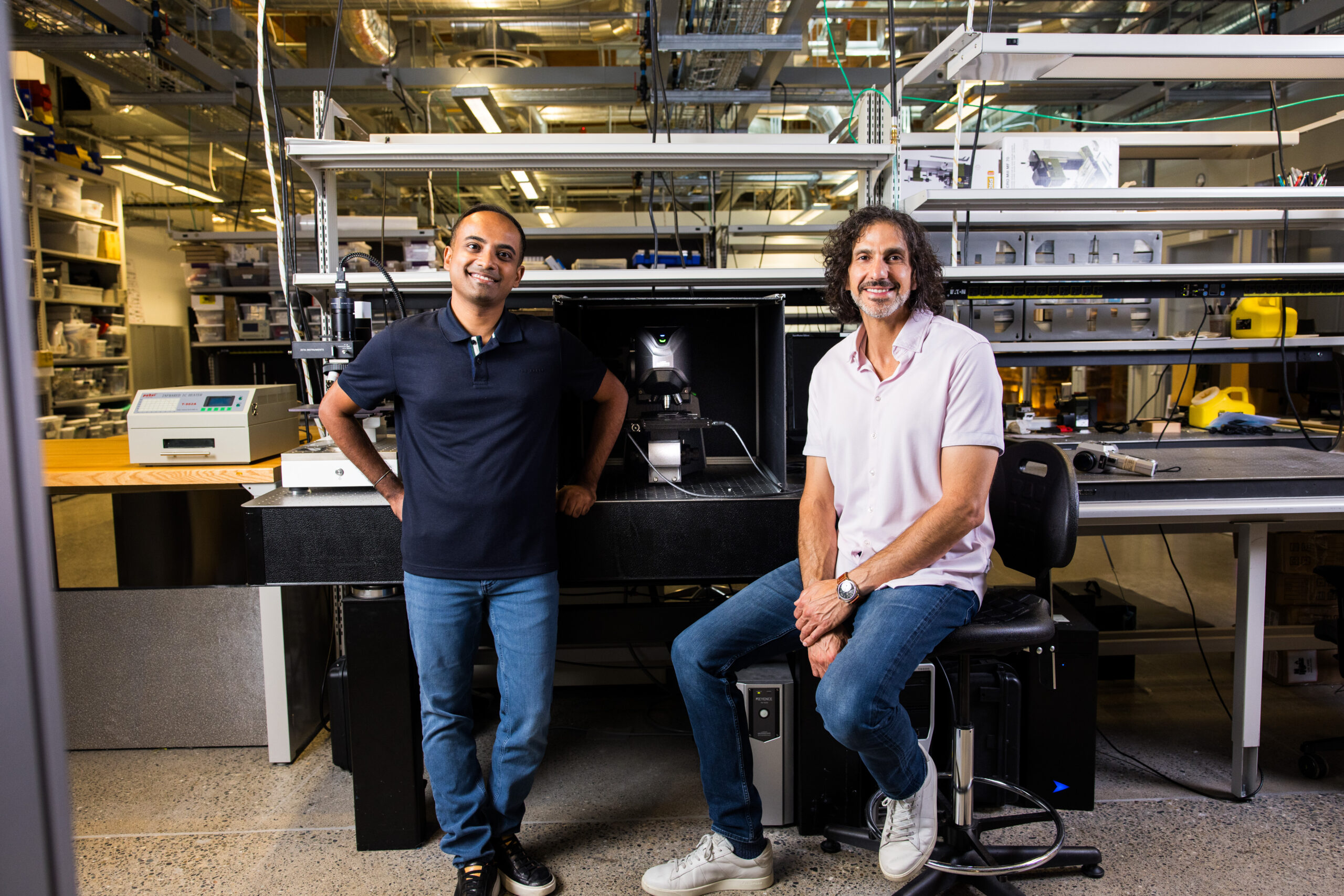 La imagen muestra a dos personas en un laboratorio o espacio industrial. Una persona está a la izquierda, con ejército de mar oscuro, jeans azules y zapatos negros. La otra persona está sentada en los taburetes correctos, con una camisa de manga corta, jeans azules, zapatillas blancas y un brazalete. Los cables se encuentran frente a una mesa negra que parece un dispositivo de los microscopios o imágenes visibles. Detrás de ellos, hay estantes que consisten en varias herramientas, equipos y contenedores, así como un techo visible con tuberías e iluminación reveladas. El piso parece ser hormigón pulido.