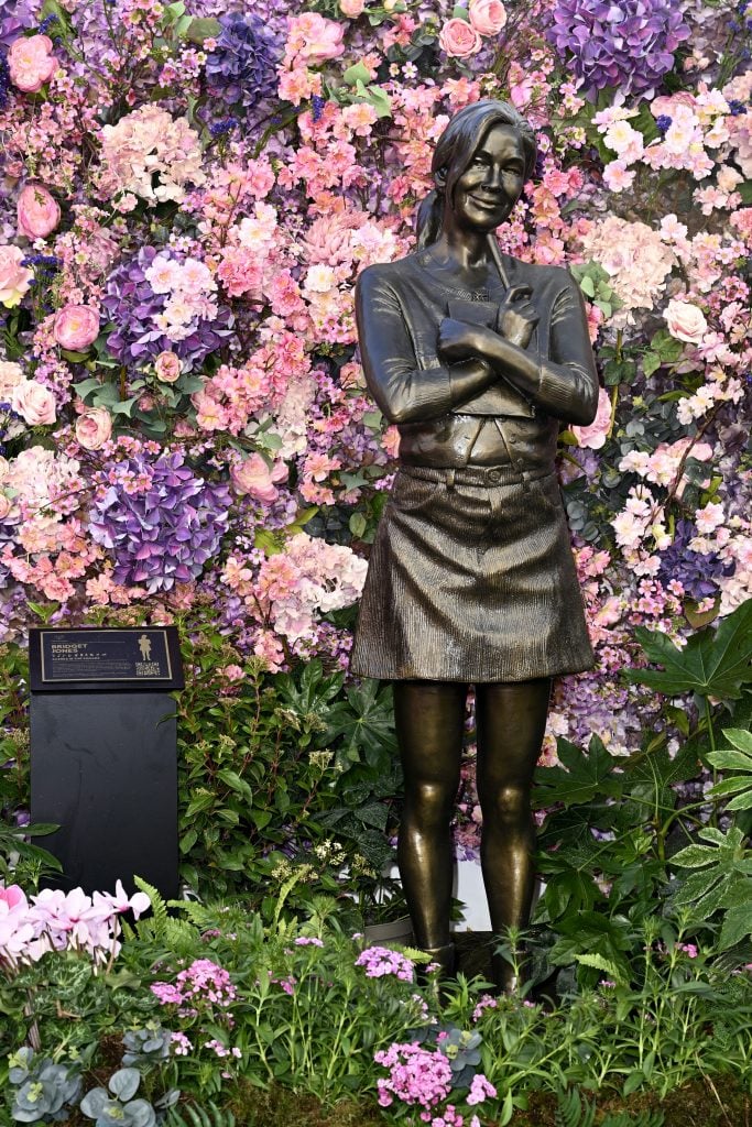 Una fotografía que muestra una escultura de bronce de Bridget Jones con un cárdigan desabrochado y una minifalda sosteniendo un bolígrafo y una libreta frente a una pared de flores de plástico de color rosa y violeta.