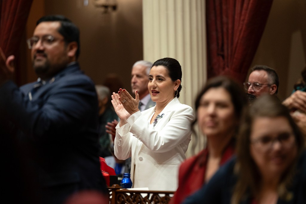 Un hombre con traje blanco aplaude mientras está dentro de una sesión legislativa mientras otros se levantan y aplauden. La escena consta de altas columnas, cortinas rojas y una cálida iluminación interior, con algunas personas en primer plano y al fondo parcialmente visibles.