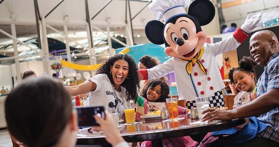 Una persona vestida con un disfraz de Mickey Mouse posa con una familia sonriente sentada en la mesa de un restaurante, disfrutando de su mágica experiencia gastronómica como titulares de la tarjeta Visa de Disney en Walt Disney World.